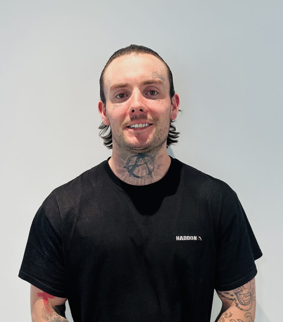 kyle a warehouse assistant at haddon equipment stands again a white wall wearing a haddon uniform shirt and smiling with his many tattoos showing