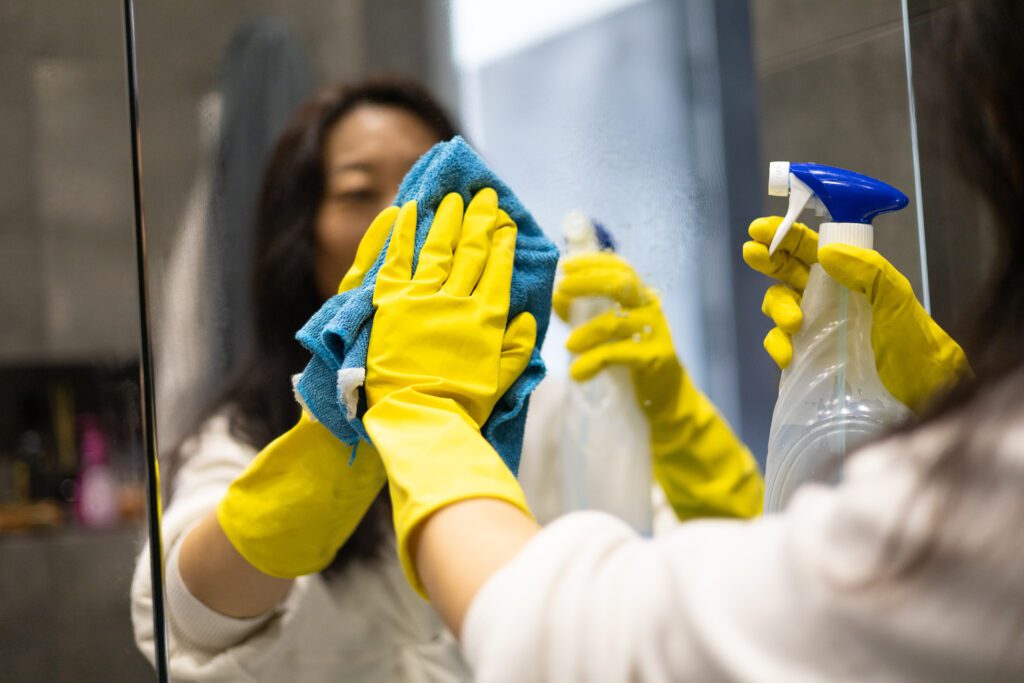 asian woman wearing rubber gloves sprays one of the best housekeeping chemicals on a hotel room mirror and wipes it with a blue microfibre cloth